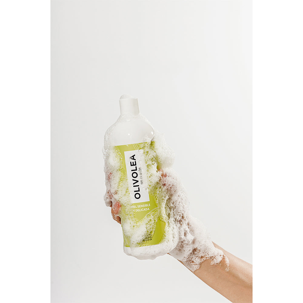 Mano sosteniendo gel de baño Olivolea en envase verde claro cubierto de espuma, fondo blanco minimalista