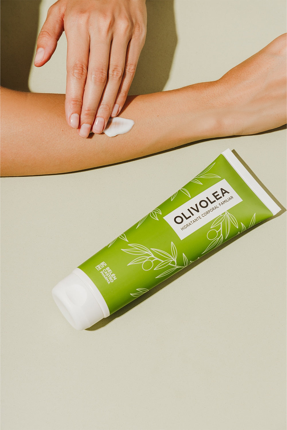 Aplicando crema hidratante corporal Olivolea en el brazo, envase verde con diseño de hojas sobre fondo beige