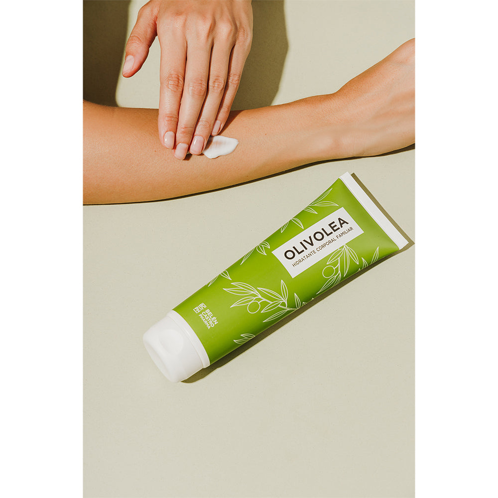 Persona aplicando crema Olivolea hidratante corporal familiar en brazo, envase verde sobre superficie beige.