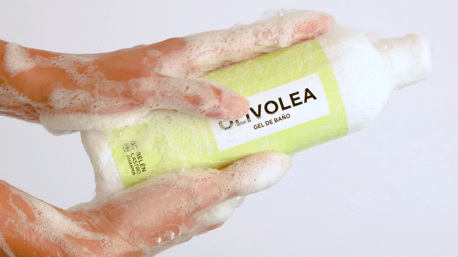 Manos enjabonadas sosteniendo botella de gel de baño Olivolea de color verde claro, con espuma cubriendo parte del envase.